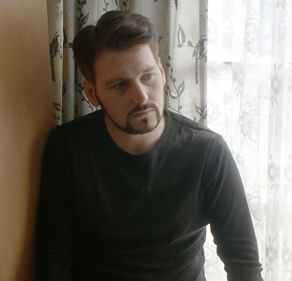 Richard Seymour has dark wavy hair, a beard and pale skin. He is wearing a black long-sleeved T-shirt and black jeans. He is seated on a windowsill against grey floral curtains looking down towards the right of the frame.