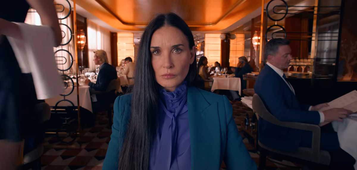 Demi Moore has long dark hair and is seated at a table in a restaurant. She is wearing a high-necked purple blouse under a blue blazer. She is staring intently across the table.