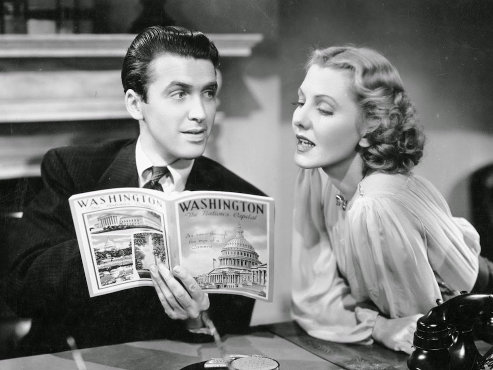 A black and white movie still portrays a man with dark gelled-back hair in a dark suit seated at a table holding a booklet about Washington, D.C. To the right of the frame, a woman with wavy blond hair and a ruffled blouse leans across the table and looks at the book mid-speech.