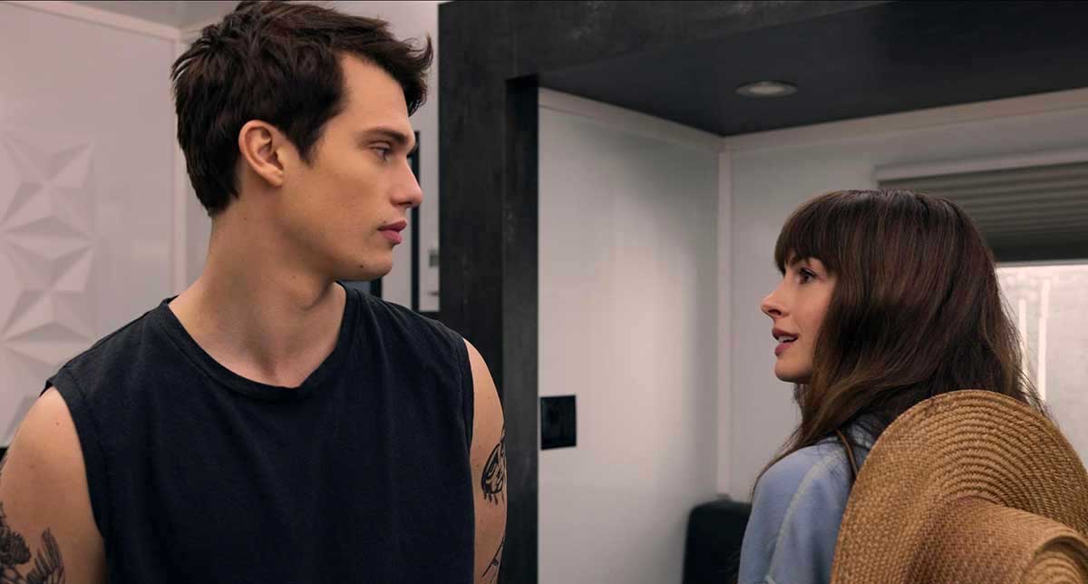 Nicholas Galitzine has short dark hair and is wearing a sleeveless black T-shirt. There are tattoos on his arm. Anne Hathaway looks towards him, mid-speech. She has long brown hair with bangs and a denim shirt. A brown straw hat hangs over her back.