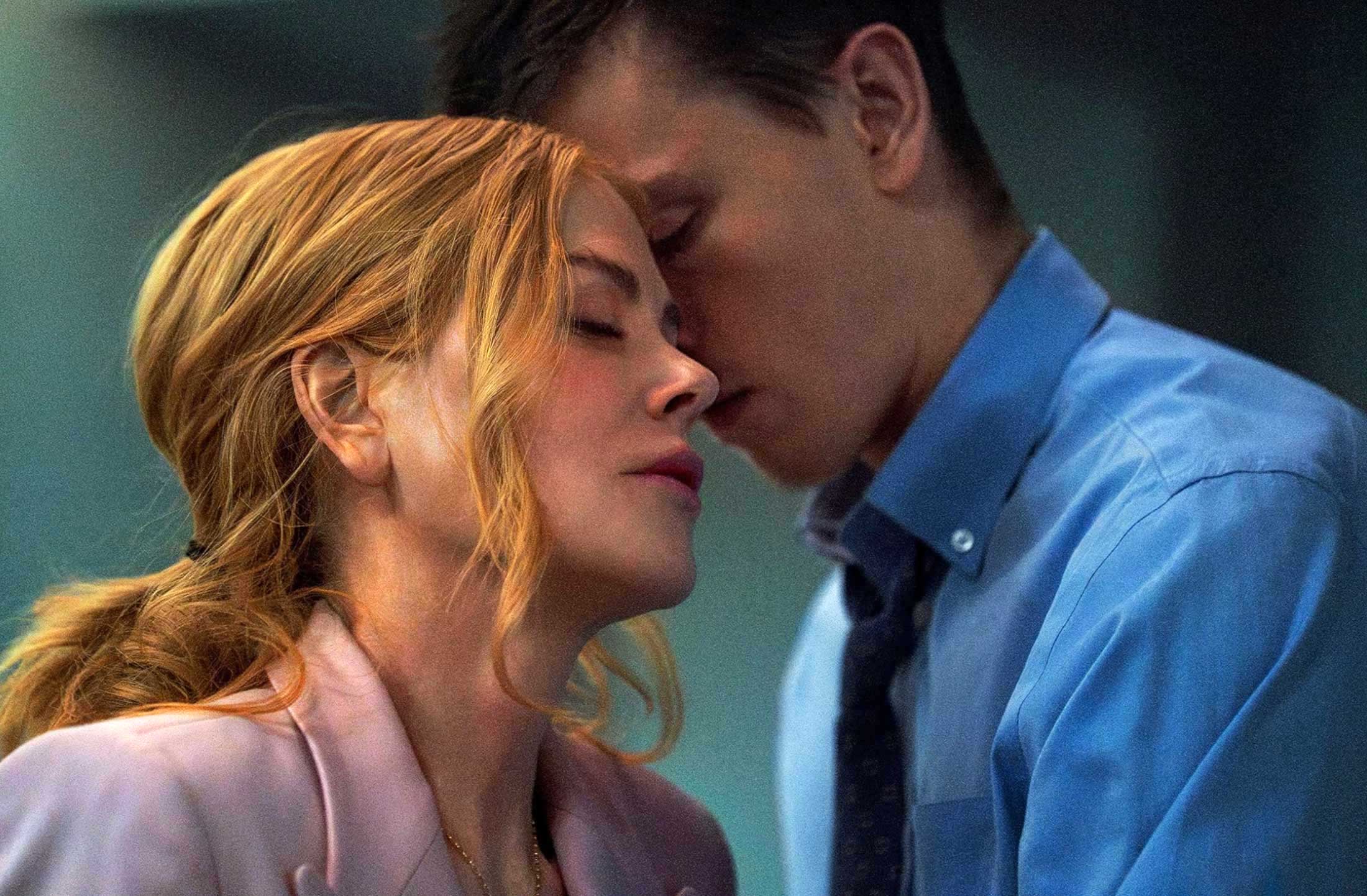Nicole Kidman has wavy blond hair and light skin. She is wearing a baby-pink blazer and is closing her eyes while leaning towards Harris Dickinson, a young man with short brown hair and a blue button-down shirt.