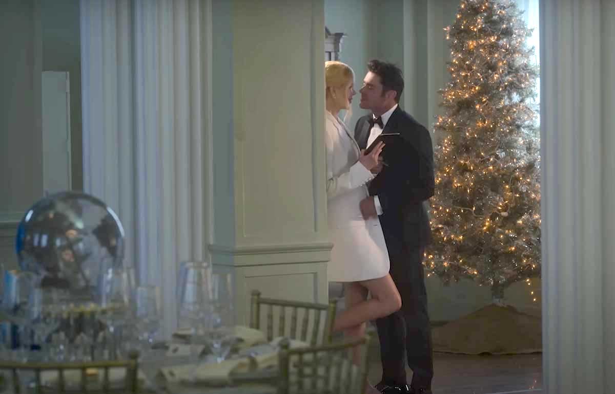 Nicole Kidman leans against a wall near a dimly lit dining room beside a Christmas tree. She has blond hair, pale skin and is wearing a cream-coloured blazer over a short skirt. Zac Efron has short dark hair and is wearing a tuxedo, moving towards her.