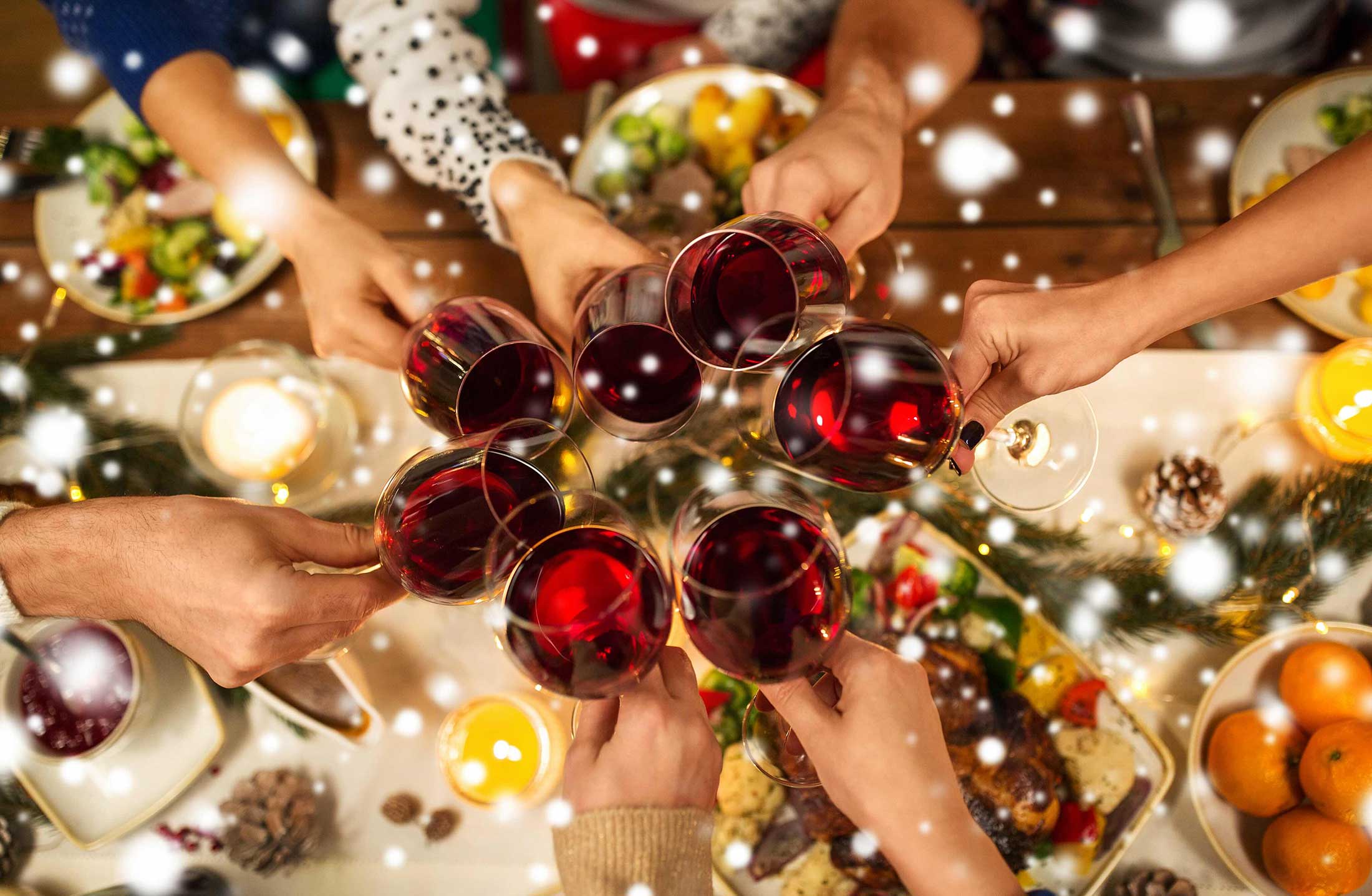 Several hands hold glasses of red wine in a gesture of 'cheers' over a sparkly, colourful table spread with food and drink for the winter holidays.