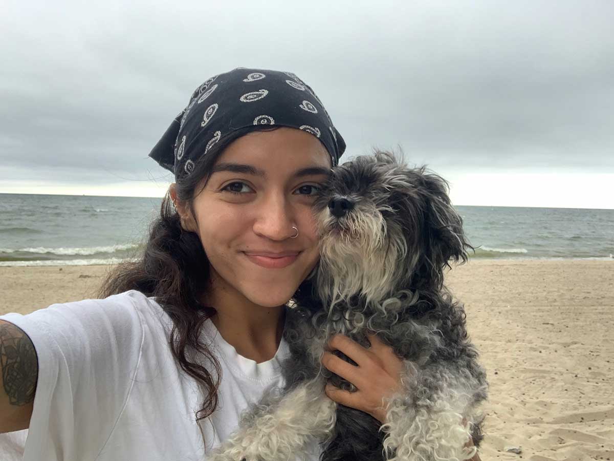 A selfie of a woman with dark wavy hair and a bandana smiles at the camera holding her dog. A beach is in the background.