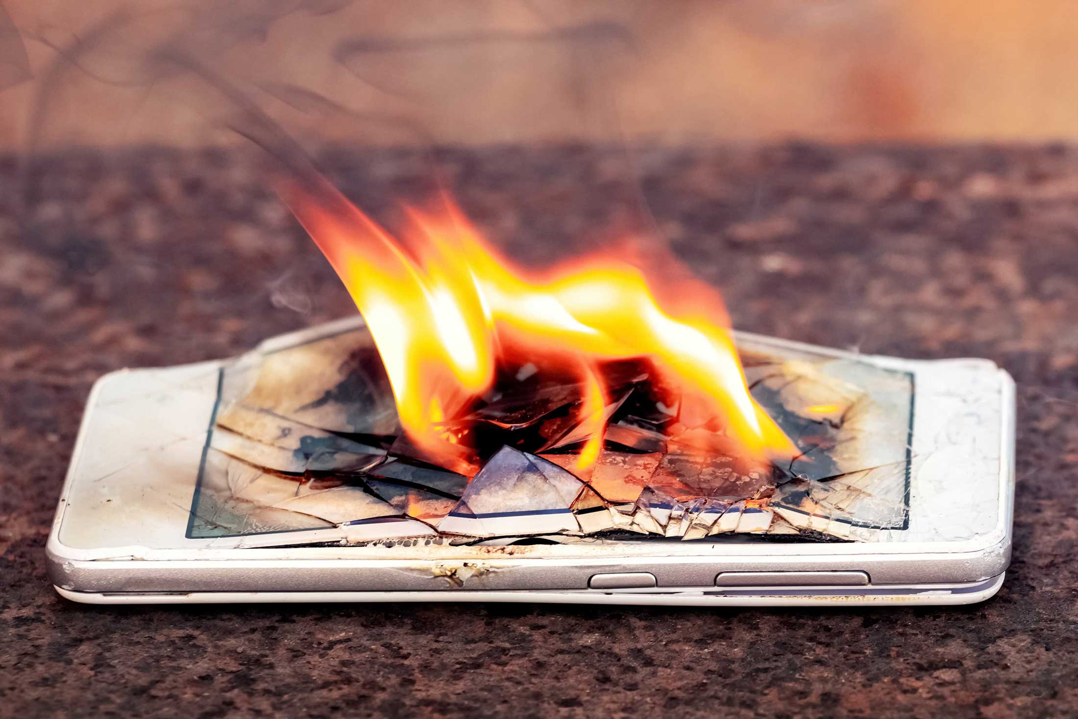 A white smartphone lies on the ground. Its screen is cracked and in flames.