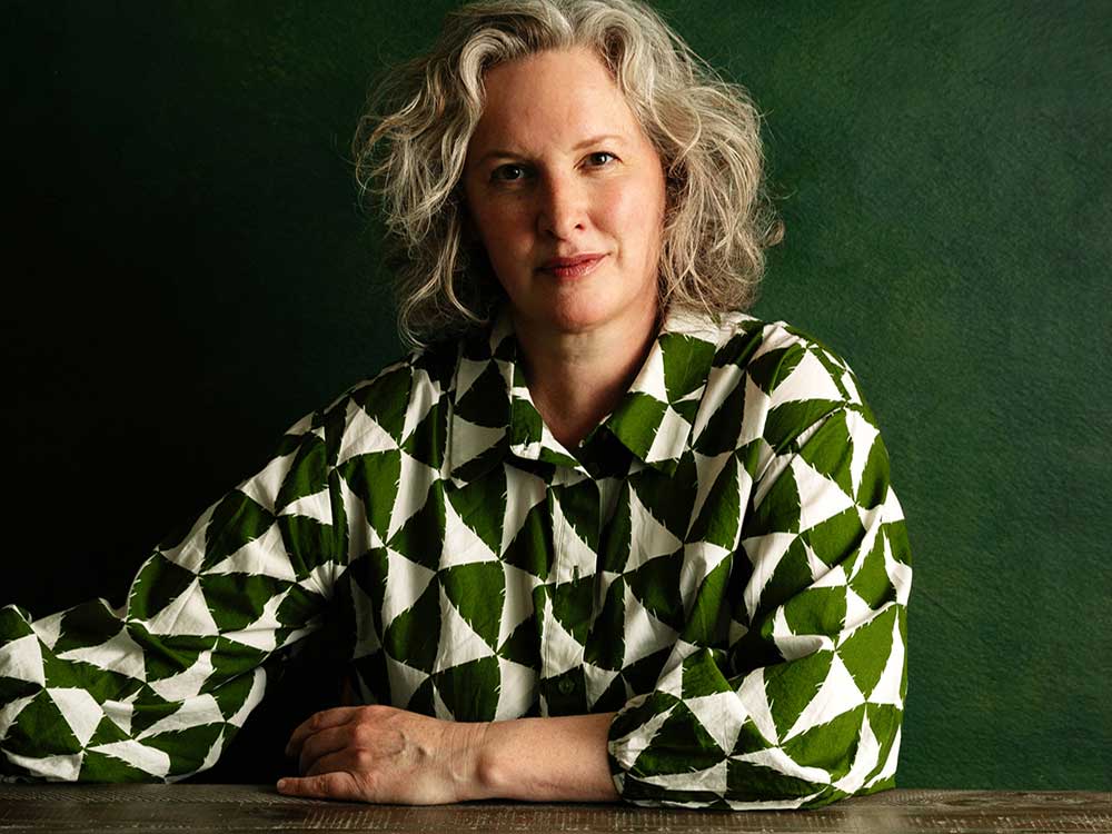 Elee Kraljii Gardiner has shoulder-length wavy grey-blond hair. She is wearing a white and green shirt with a geometric pattern. She is seated at a wooden table against a dark green background.