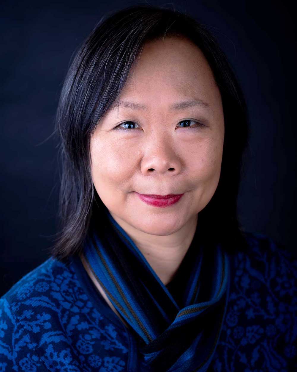 Fiona Tinwei Lam has shoulder-length black hair. She is smiling gently at the camera and wearing a blue floral shirt with a blue scarf. She is photographed against a dark blue background.