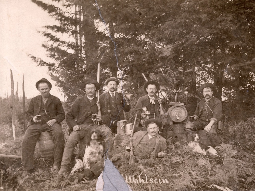 An archival sepia photograph depicts a group of pioneer men posing together in a forested setting. They are wearing dark clothing and hats, and some are holding rifles and mugs of beer. A dog is seated in front of the men to the left of the frame.