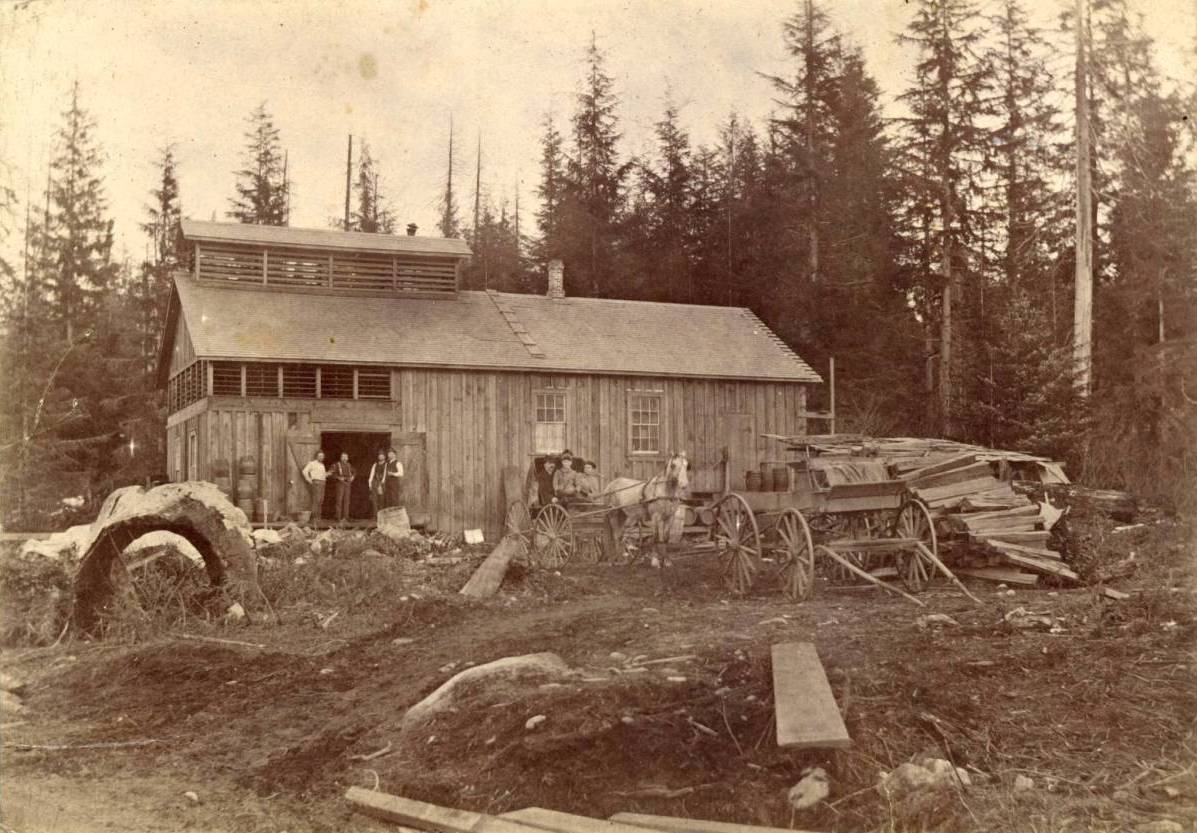 An archival sepia photo of a wooden structure in a wooded area that housed the Columbia Brewery in Vancouver&rsquo;s earliest years. A horse-drawn carriage is in front of the building and four men are standing in the doorway.