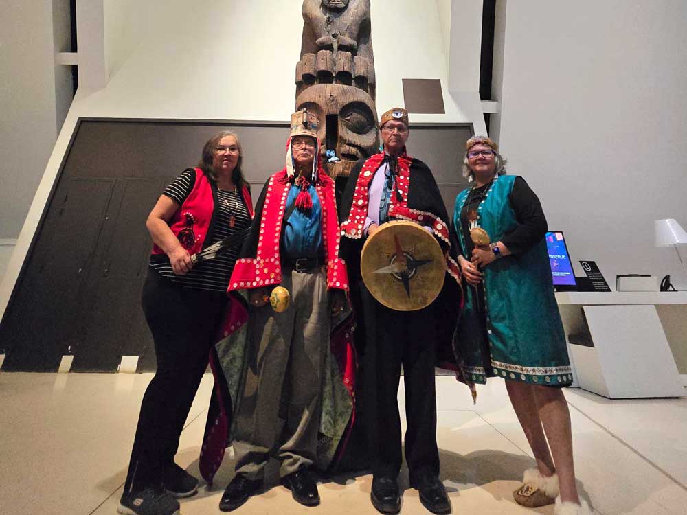 Four people, two men and two women, stand in front of a large, carved wooden object. They are wearing traditional Indigenous regalia.