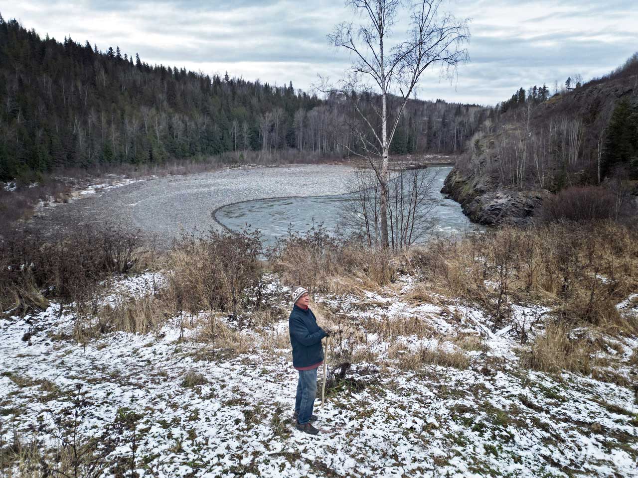 A man wearing a toque and winter coat, and leaning on a wooden staff, stands in a grassy area. Behind him there is a river and forests.