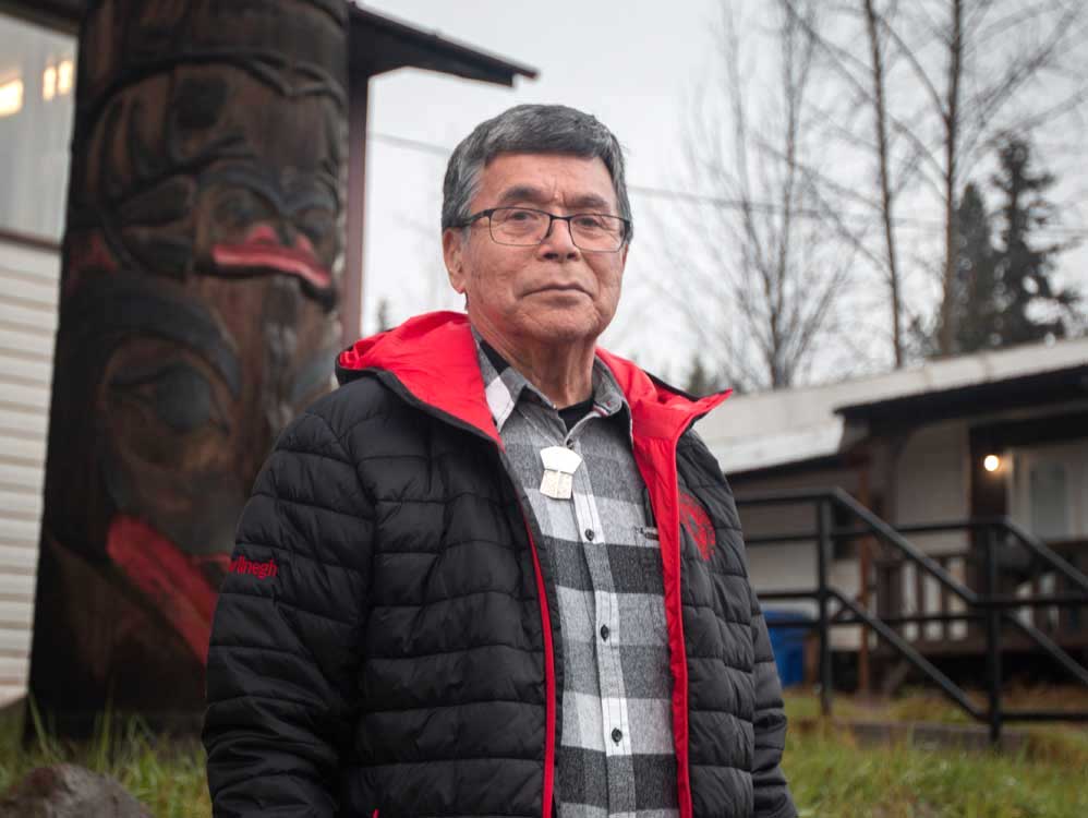 An older man with greying hair, wearing glasses and a winter coat, stands outdoors looking at the camera. Behind him are buildings and a large wooden carving.