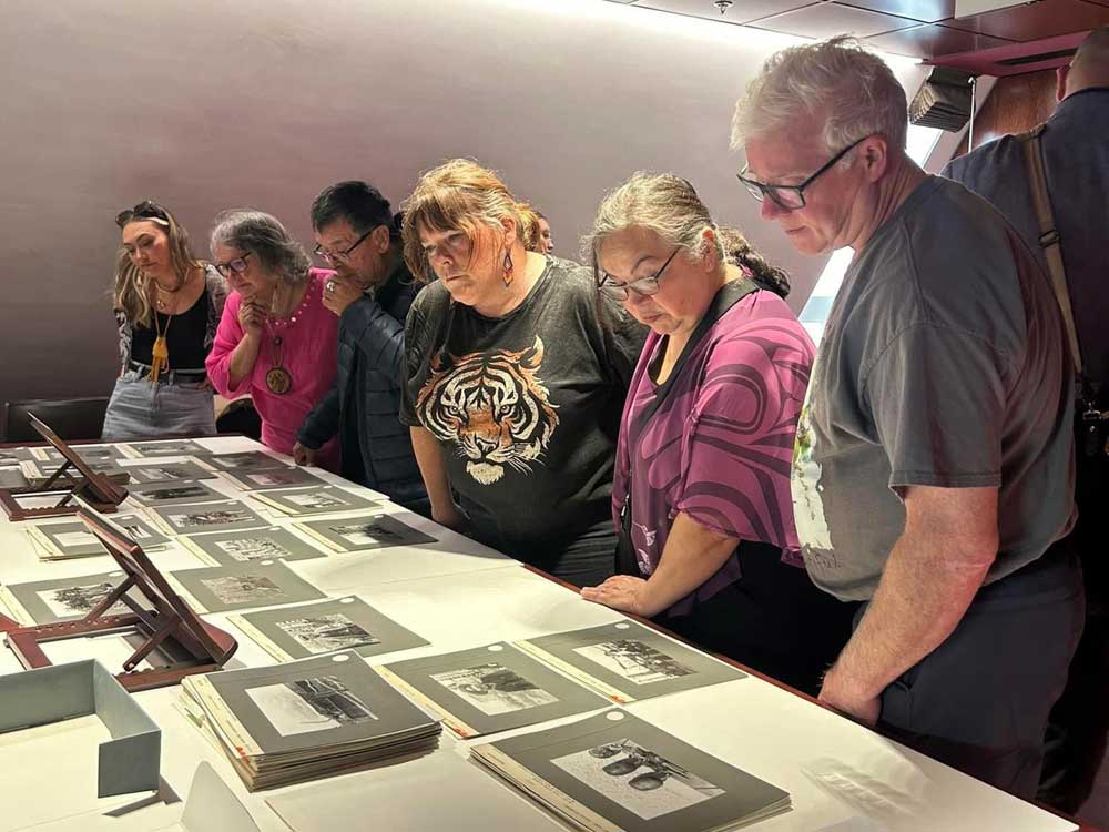 Six people stand in a row examining black and white photos that are laid out on a white table.