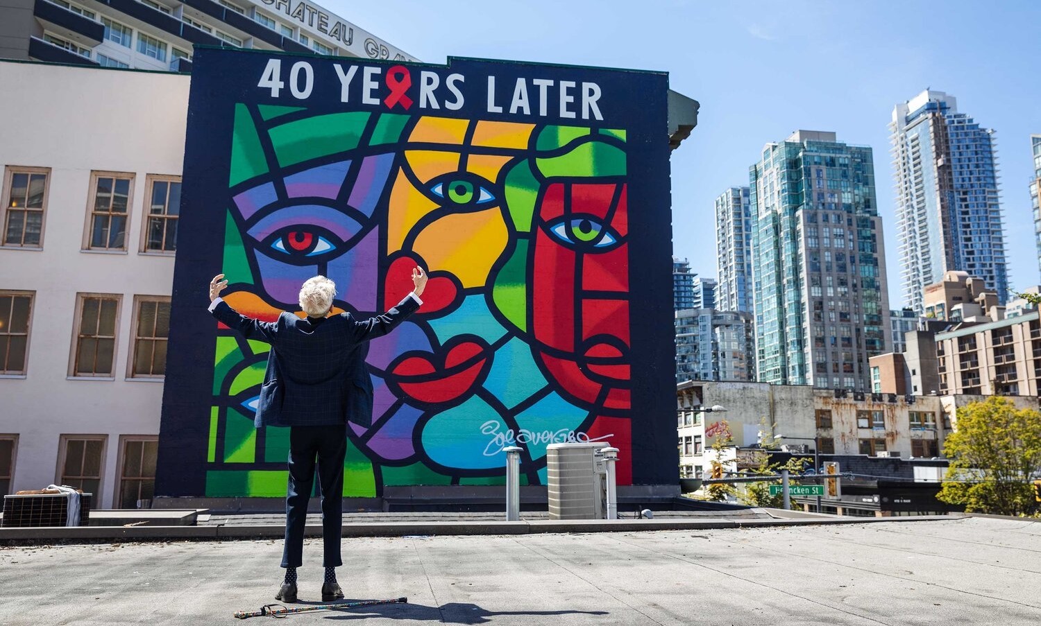 A thin man with grey hair wearing a dark suit stands with his back to the camera and his arms outstretched as he faces a colourful mural on the side of a building.