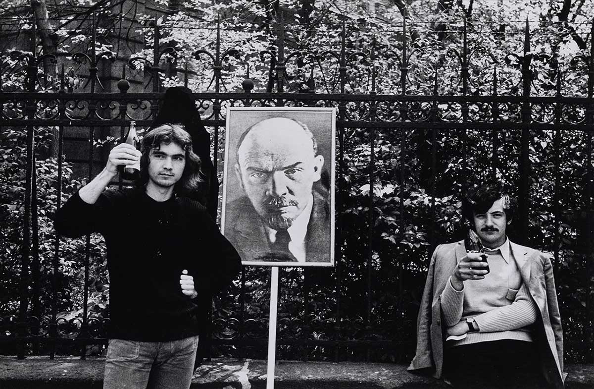 A black and white photograph depicts two young men outdoors against a garden wall, each holding a bottle of beer. Between them is a photograph of Vladimir Lenin on a wooden stick.