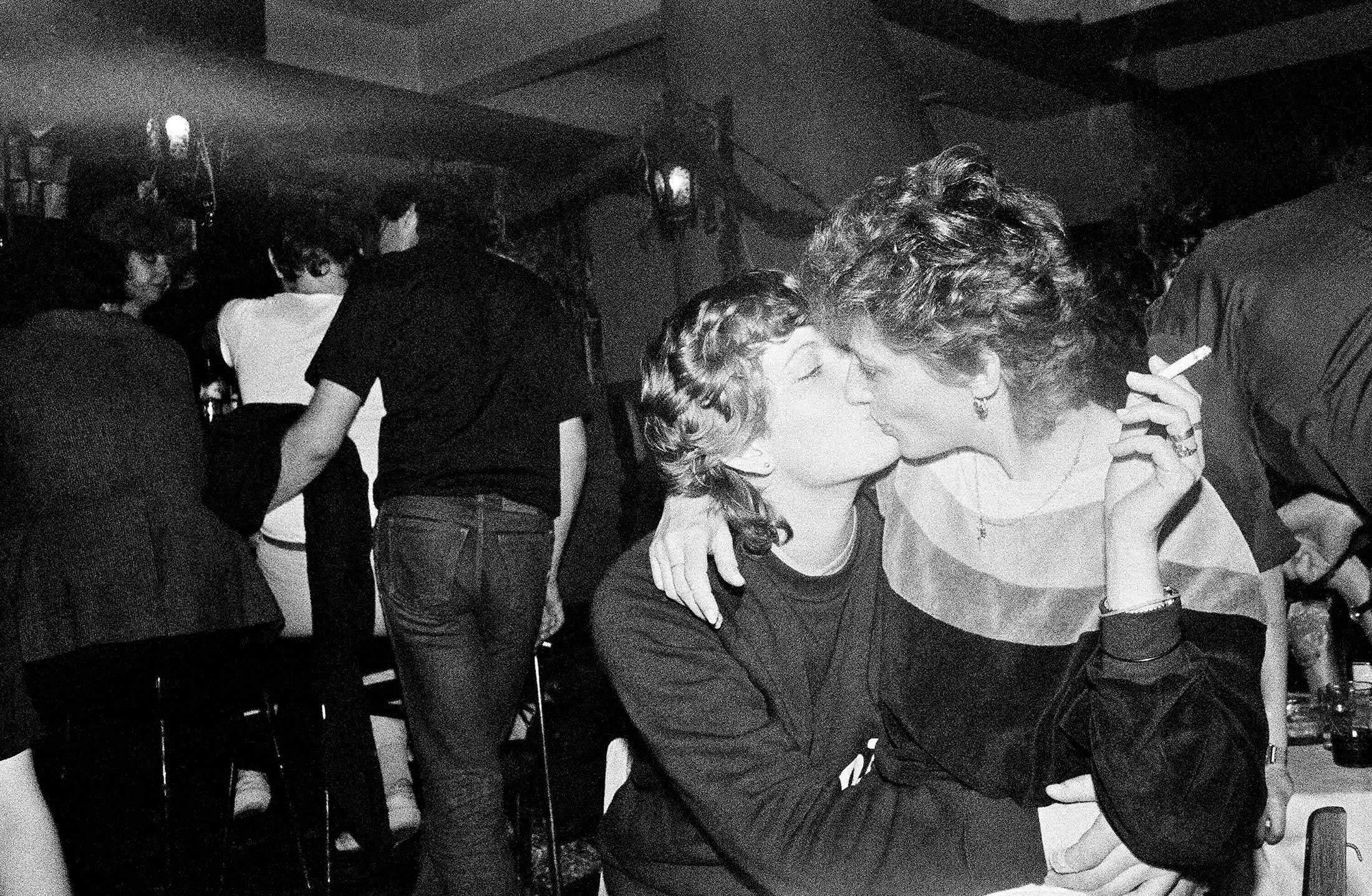 A black and white photograph of a nightclub interior. In the foreground to the right of the frame, two people with short blond wavy hair are kissing. The person on the right is holding a cigarette.