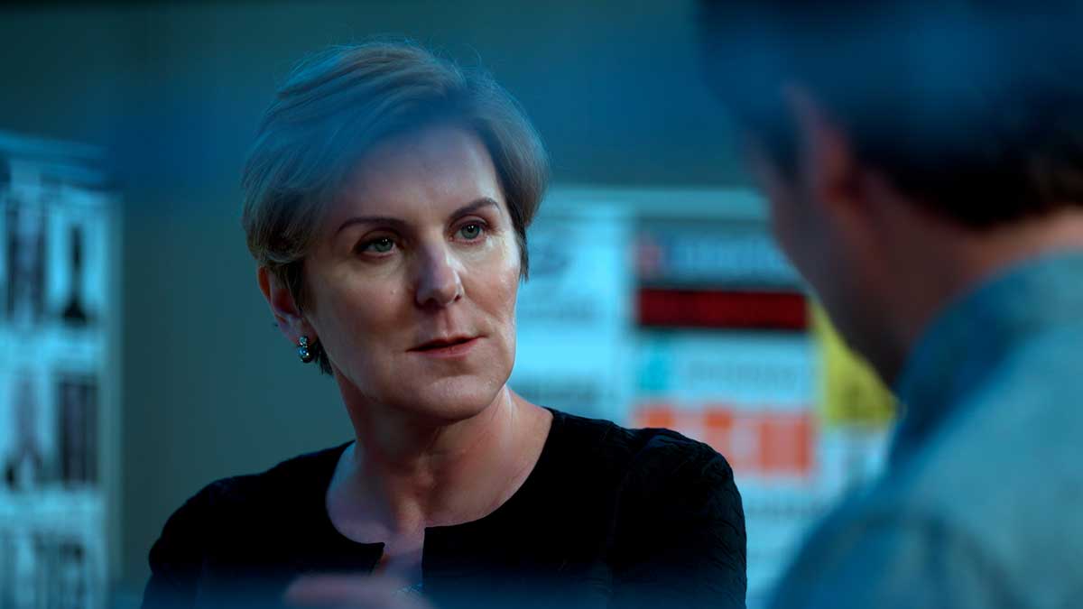 Alexandra Wrage has short blond hair. She is wearing a dark top and speaking to a person across from her who is in soft focus with their back to the camera. She is in a darkened interior space under blue light.