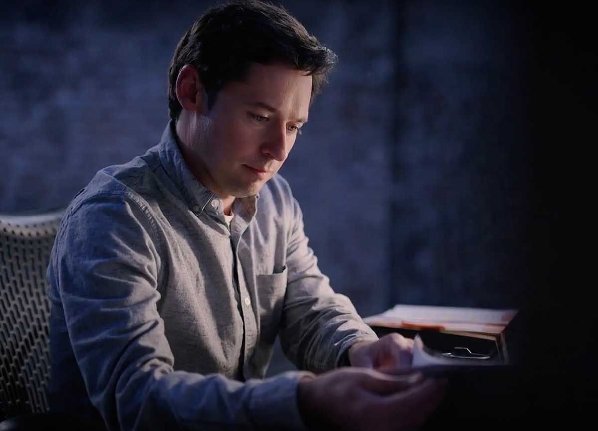 Nick McKenzie has short brown hair and is wearing a light blue button-down shirt. He is seated at a desk in a darkened room, examining a stack of documents.