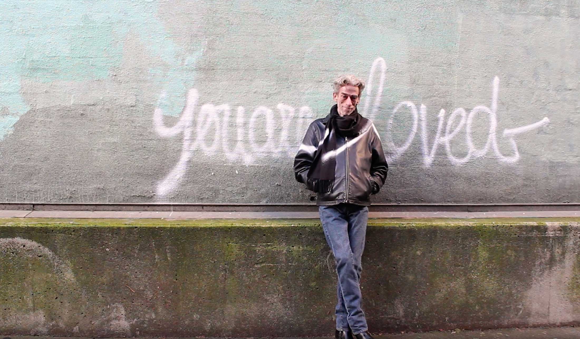 Joe Average has wavy greying hair. He is wearing a black leather jacket, a thick black scarf, blue jeans and black shoes. He is leaning against a concrete wall. The phrase “You are loved” is spray painted in baby pink cursive across the wall and across Average’s jacket.