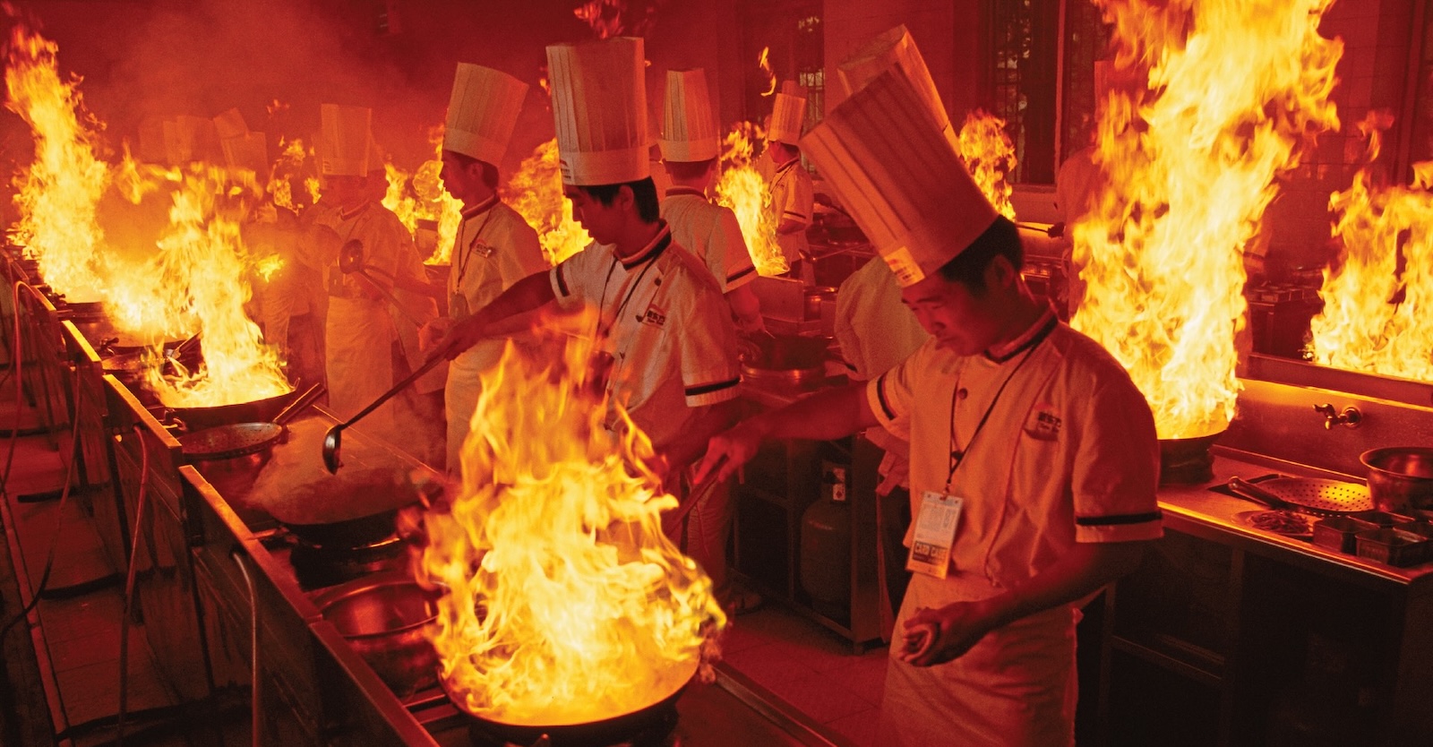 People in white chef’s uniforms and tall chef’s hats stand in two rows, holding long ladles before woks full of fire; the area around them is red and orange, reflecting the flames.