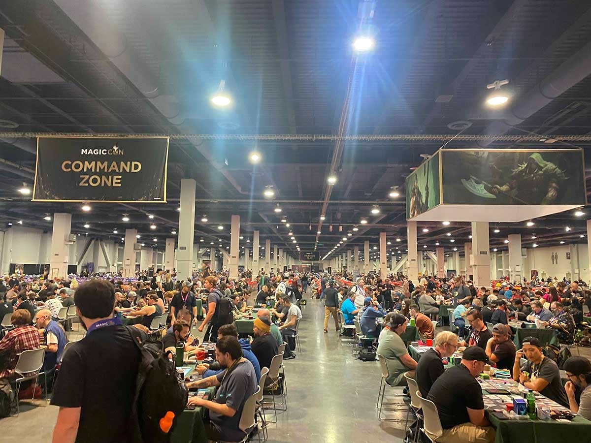 A busy interior space is set up with long rows of tables at which people are seated playing Magic: The Gathering. The high black ceilings carry bright white lights. A high black banner to the left of the frame reads 'MagicCon Command Zone.'