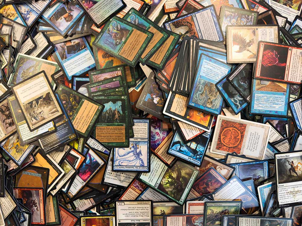 A close-up photograph of hundreds of colourful Magic: The Gathering playing cards stacked on each other across a flat surface.