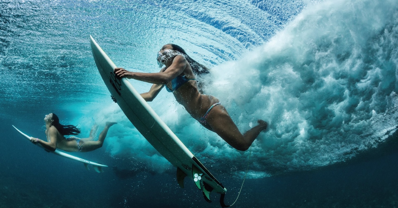 Two people swim underwater, holding white surfboards below their bodies. They both have long dark hair and bubbles are visible near their noses and mouths.