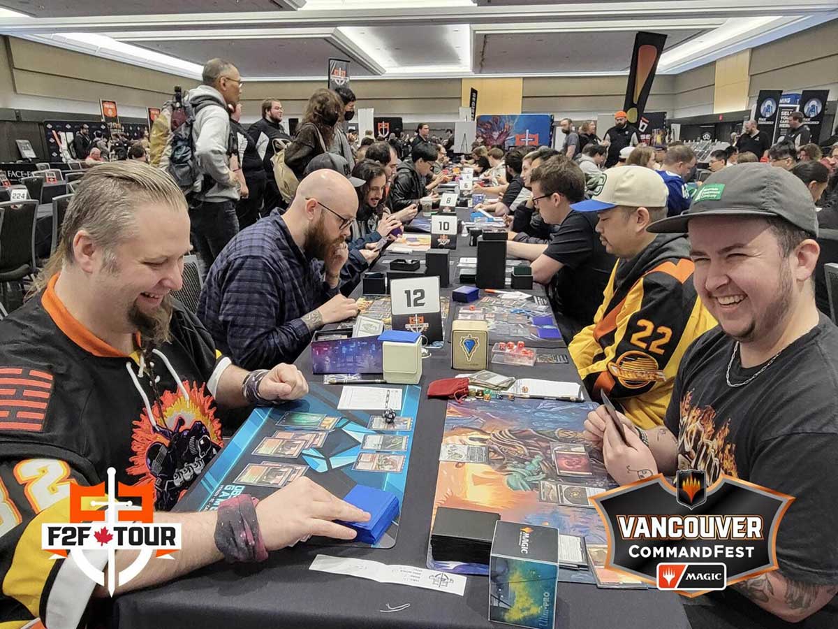 Many people are seated across from each other at a long row of tables fitted with black tablecloths. They are playing the fantastical card game Magic: The Gathering at a local competition. Mel Woods, the author, is smiling at the camera to the right of the frame. They are wearing a dark grey ball cap, a black graphic T-shirt and a silver necklace.