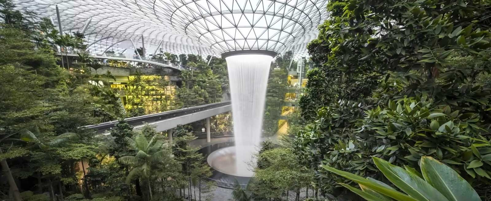 The white ceiling of an arboretum opens to a large funnel of water flowing down into a circular concrete pit below. Around it are lush green indoor vegetation and grey futuristic walkways.