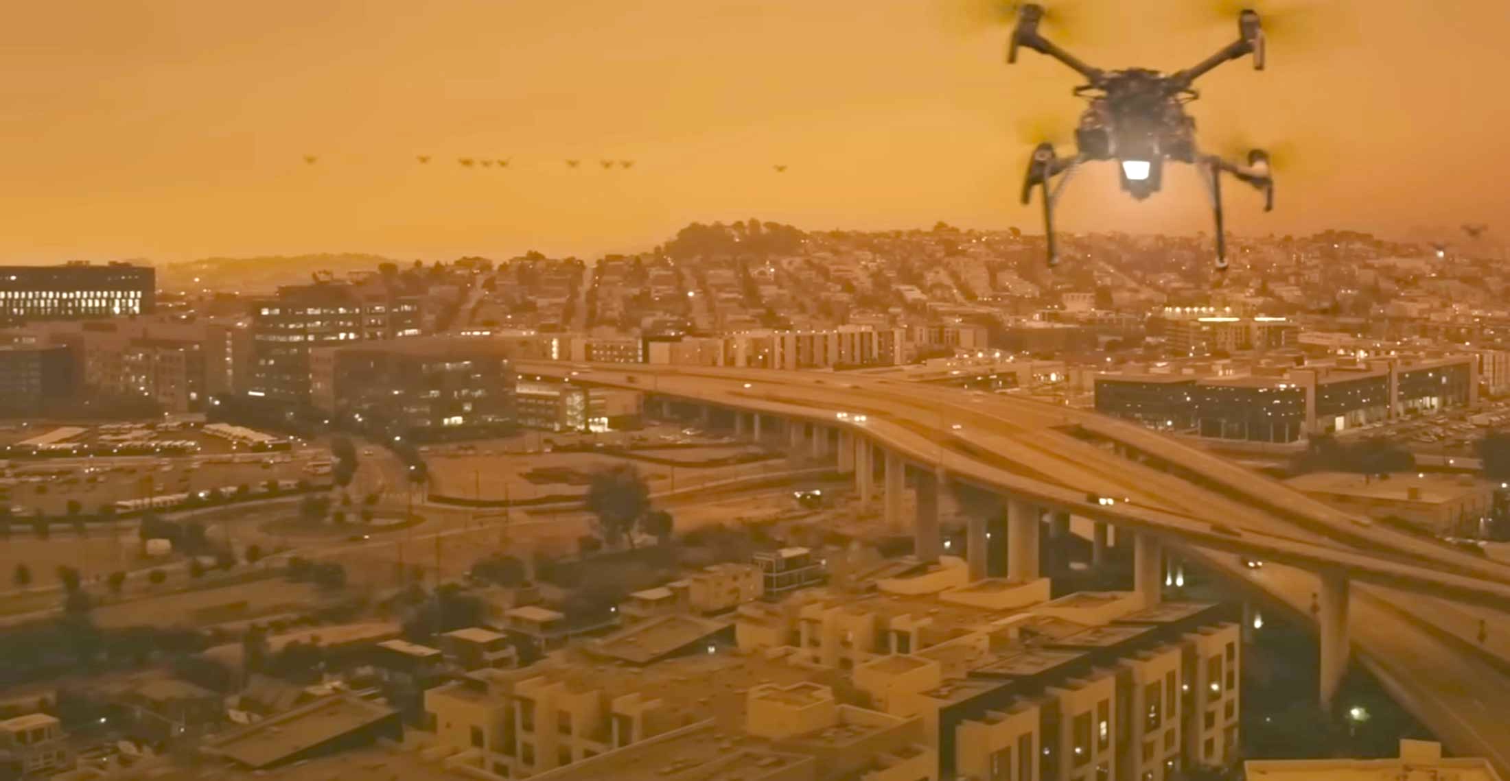 A drone flies above the elevated motorways of a busy metropolis. The sky is orange and hazy.
