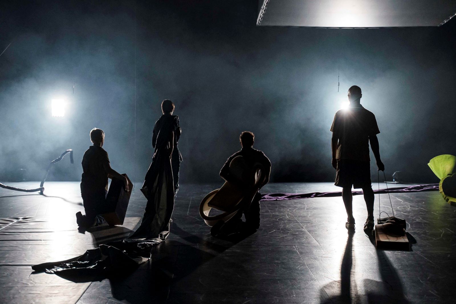 Four performers stand with their backs to the camera on a darkened stage, holding an assortment of large fabrics and props. White lights shine at them, illuminating them in silhouette.