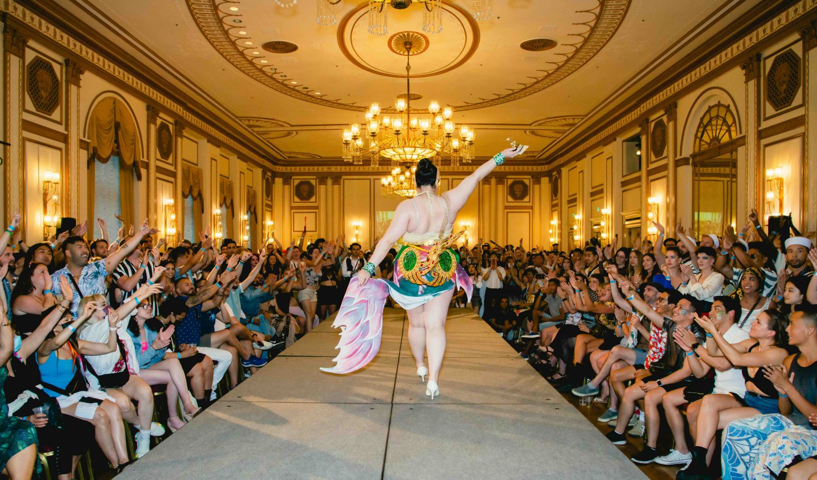 A person in a dark updo, backless dress and flesh-coloured high heels walks down a runway, their right arm raised to the crowd. Their left hand holds a pink wing-like train of their jewel-toned skirt. They are walking towards a chandelier and a crowded audience of young adults in casual streetwear raises their arms aloft in applause on either side of the runway.