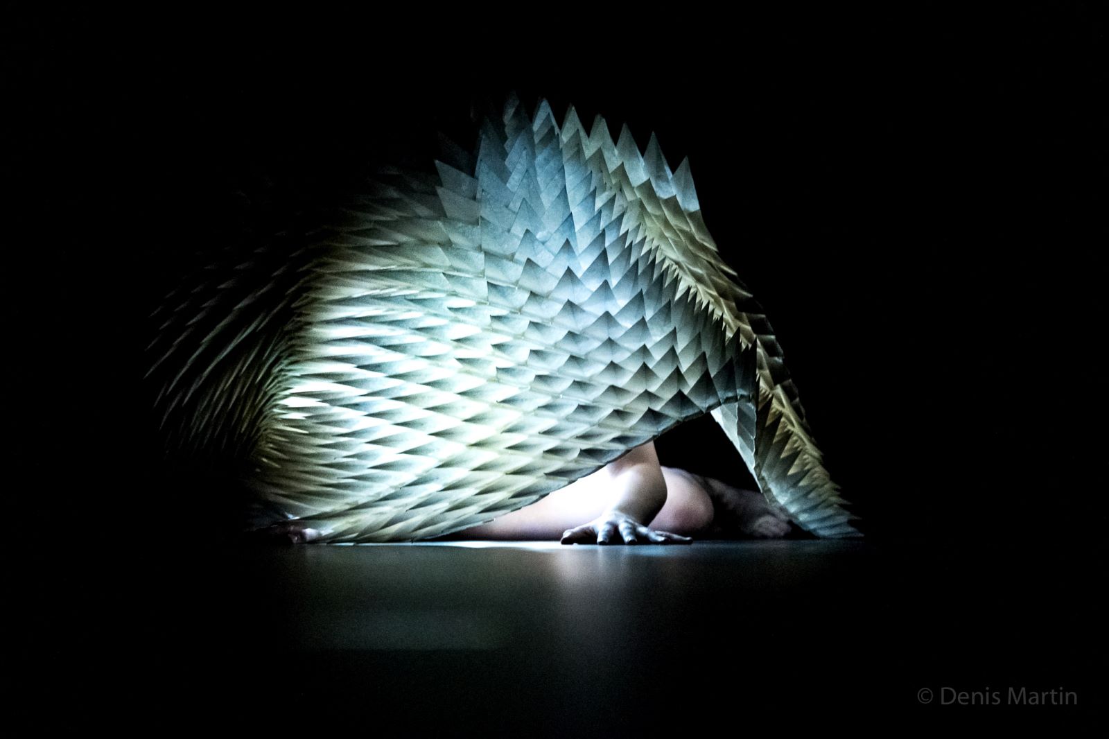 The bare arm and leg of an actor is visible beneath an illuminated fabric cover in seafoam that resembles the scales of a dragon or large reptile. They are on the floor of a stage against a black background.
