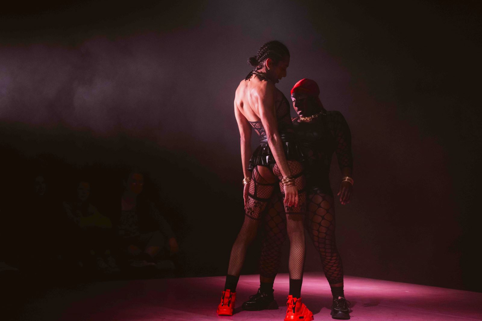 Two actors face each other on a darkened stage. The person in the foreground wears a black corset, fetish gear, fishnets and red high-top sneakers. The person facing them is in partial darkness, similarly attired.