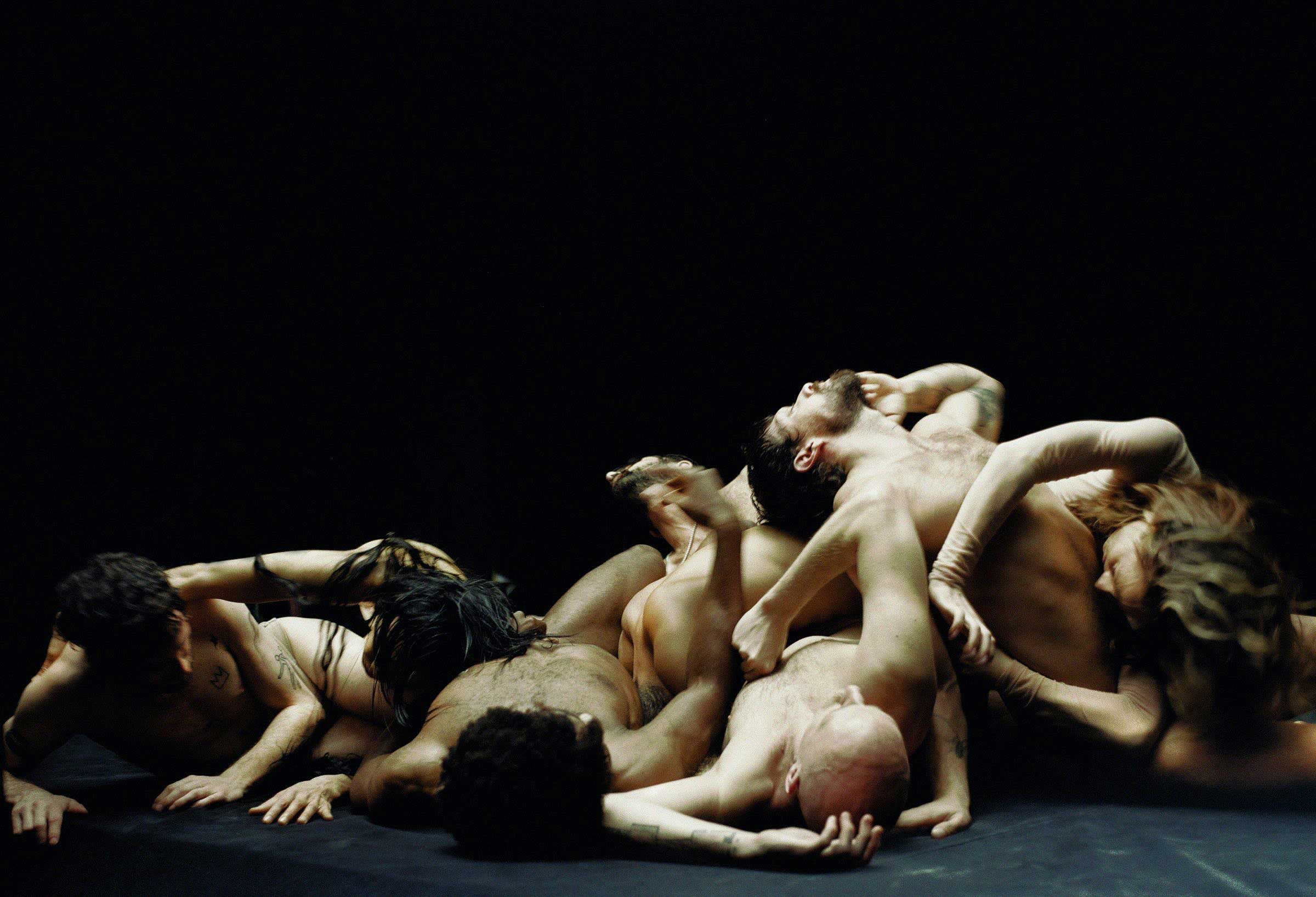 An assembly of bare-chested actors move together across a dark stage floor against a black background.