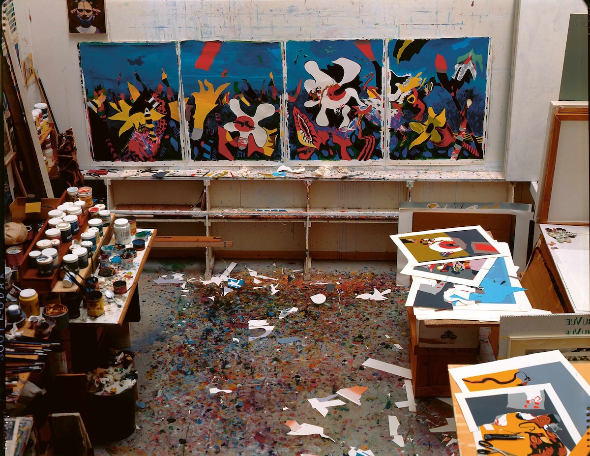 A photograph of an art studio features trays of paints and brushes on the left, a wall of paintings in progress in the middle and assorted prints on the right.