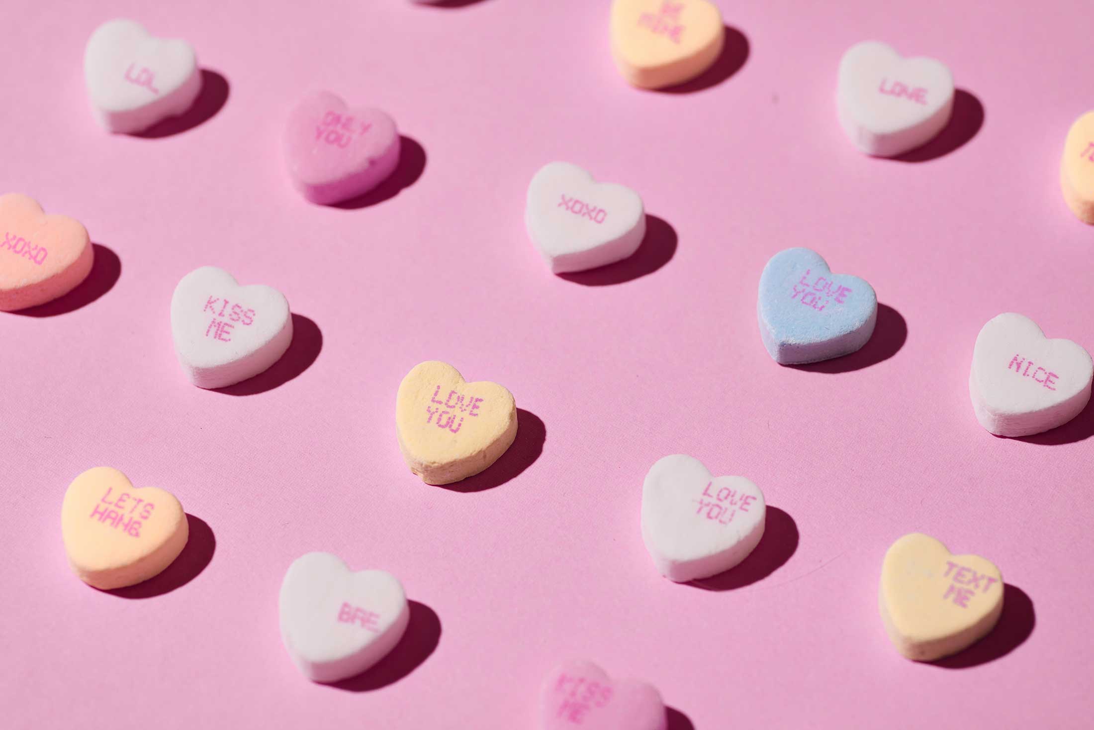 Pastel-coloured candy hearts are laid flat against a pink background and shot from above with bright lighting. The hearts are inscribed with messages like 'Love you' and 'xoxo.'
