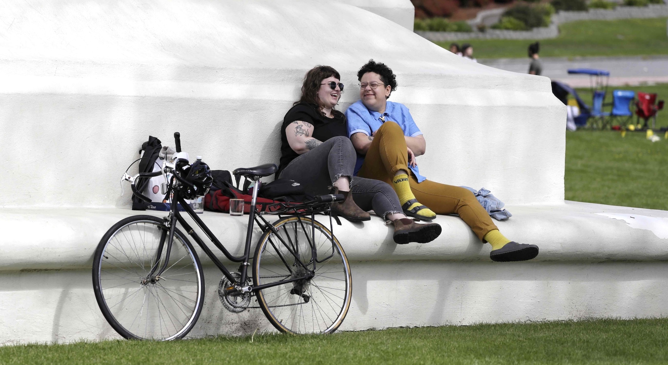 A bicycle leans against the base of a large white marble monument upon which two people sit together on its lower ledge. Hannah Smith, left, has shoulder-length dark hair, pale skin, sunglasses and a black T-shirt with dark grey jeans. Jabree Robinson, right, has short dark curly hair, medium-light skin, a short-sleeved blue shirt and tan trousers. It’s a spring day and people are gathering with lawn chairs in the background.