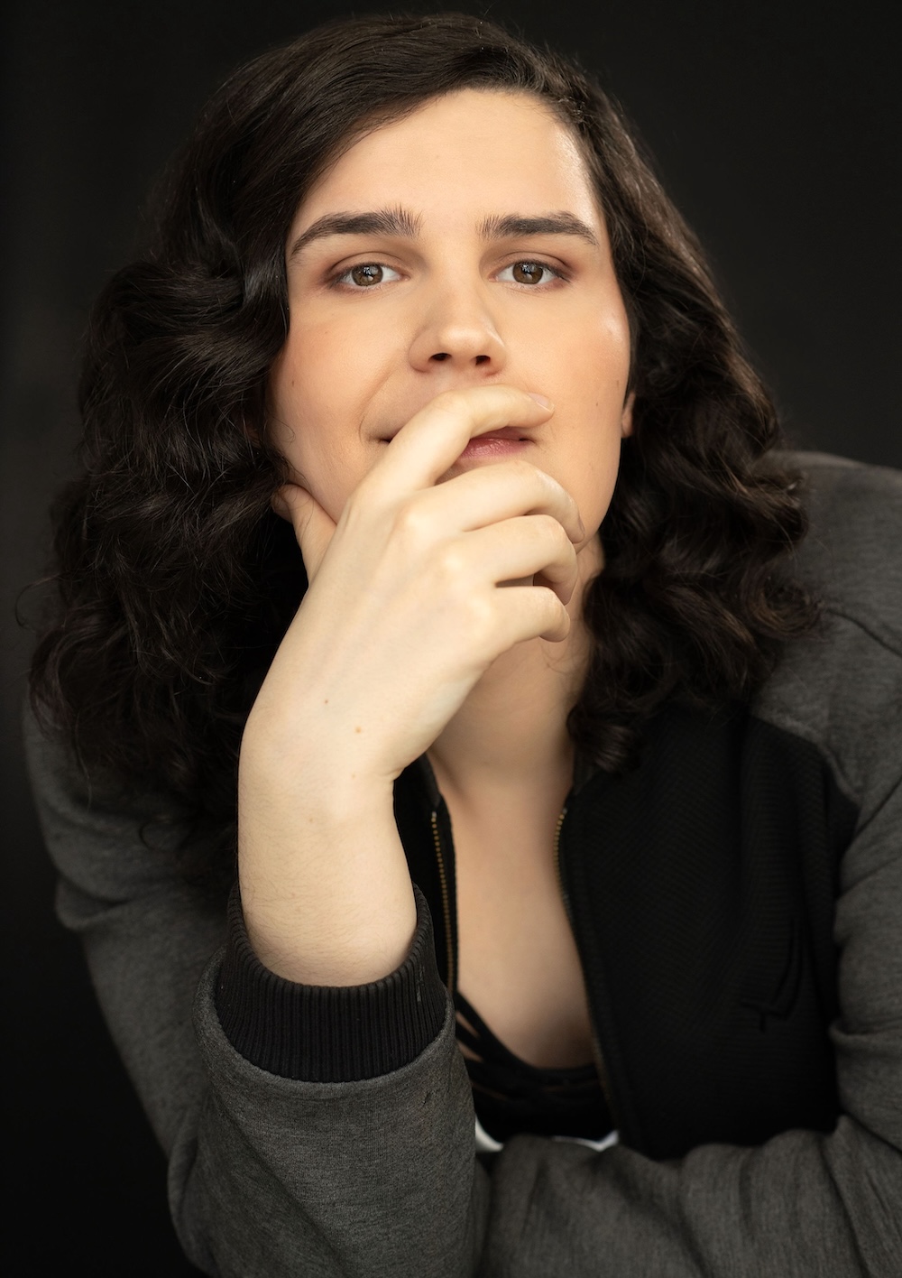 Eden Kaminski has long wavy dark hair and is wearing a grey jacket. She is leaning forward and resting her chin in her hand against a black background.