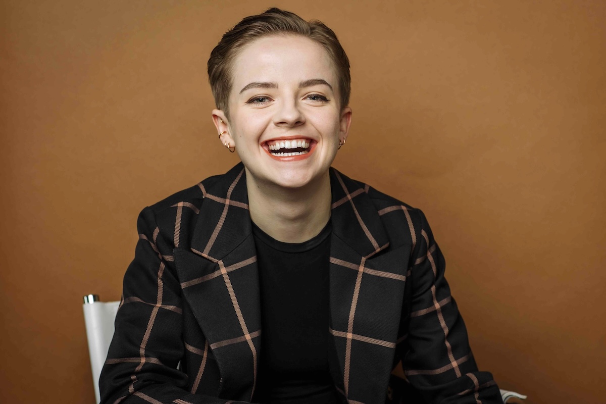 Erin Purghart has short brown hair and is wearing a black blazer with wide, light brown, checkerboard-style lines. They are seated against a copper-coloured background. They&rsquo;re looking at the camera and laughing.