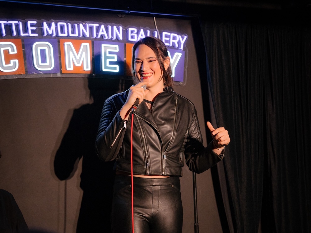 Kay has long dark hair pulled back in an updo. She is standing on a small comedy stage in the Little Mountain Gallery in Vancouver; a neon sign that reads 'Little Mountain Gallery Comedy' is on the black wall behind her. Kay is wearing a cropped black leather jacket and black pants. She is holding a microphone on a black stand and smiling.