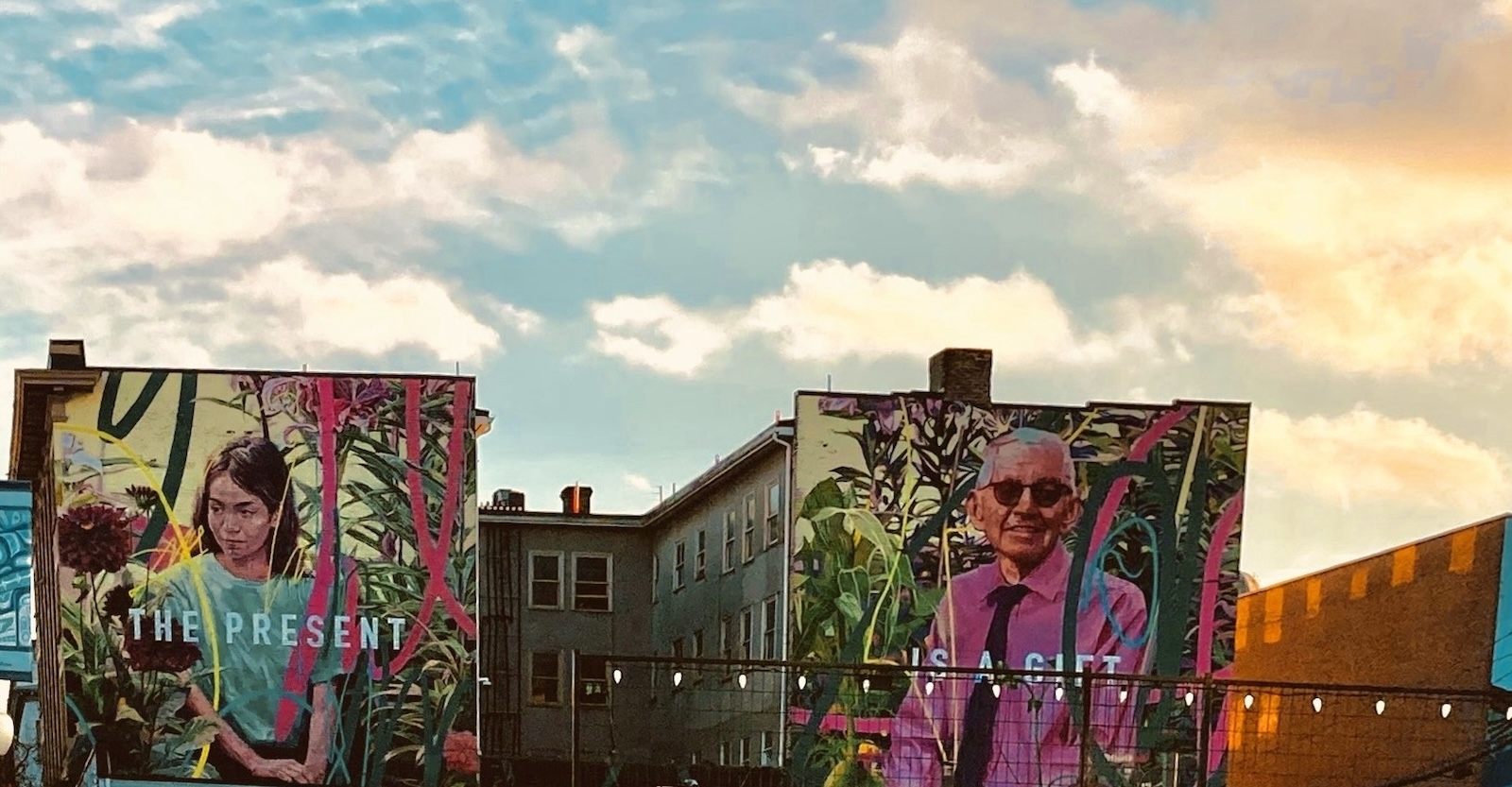 A mural stands across two highrise apartment buildings against a bright sky. On the left is a painting of a girl with brown hair and a green shirt; on the right is a painting of an older man with grey hair, glasses and a pink shirt. The girl&rsquo;s panel reads 'the present' and the man&rsquo;s panel reads 'is a gift.'