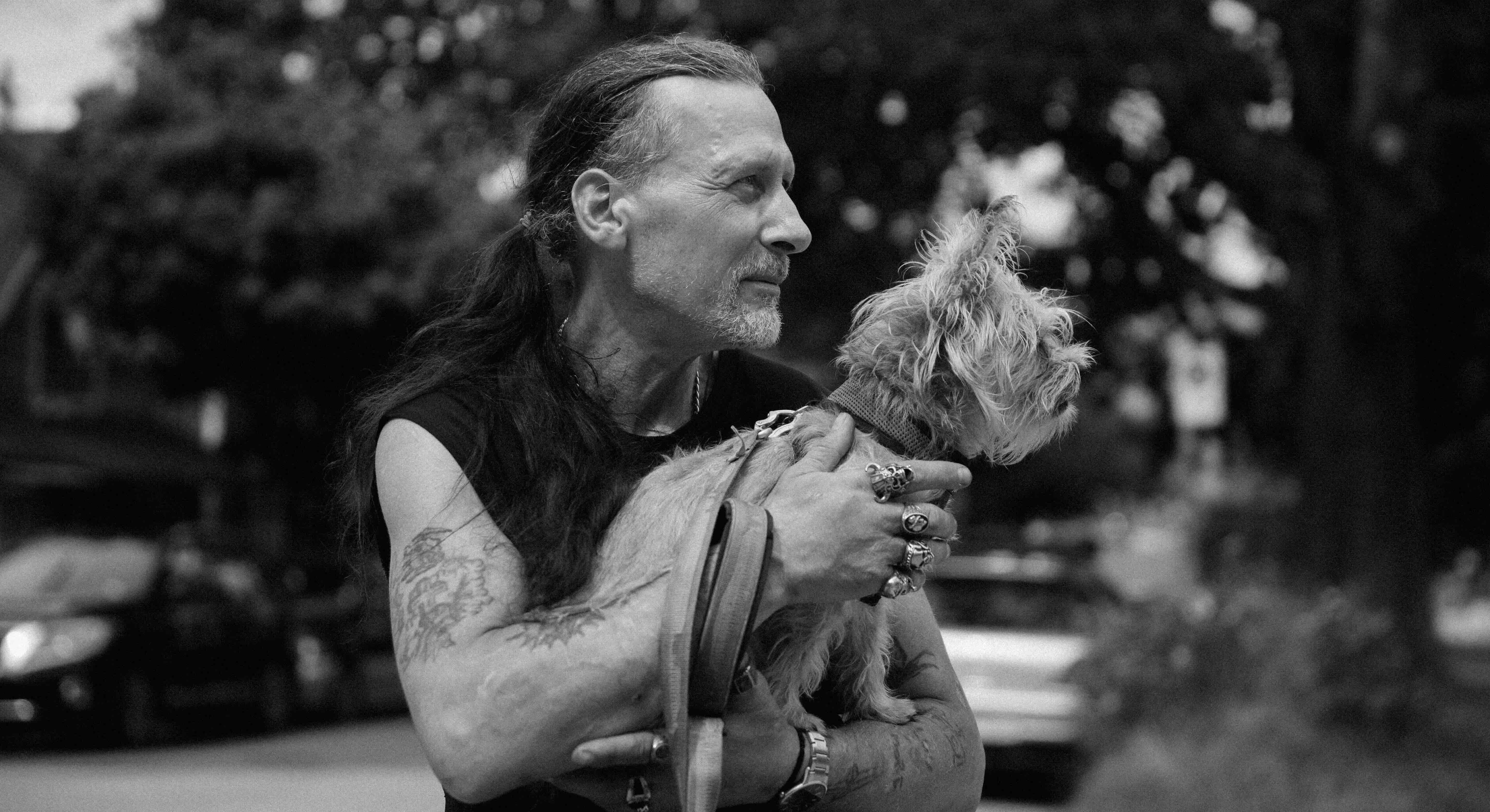 A black-and-white photograph features Martin Steward has long hair tied back. He is looking towards the right of the frame and wearing a sleeveless black shirt. He is holding a small dog in his arms who is looking in the same direction as Steward. They are standing on a residential street with leafy trees and parked cars in the background in soft focus.