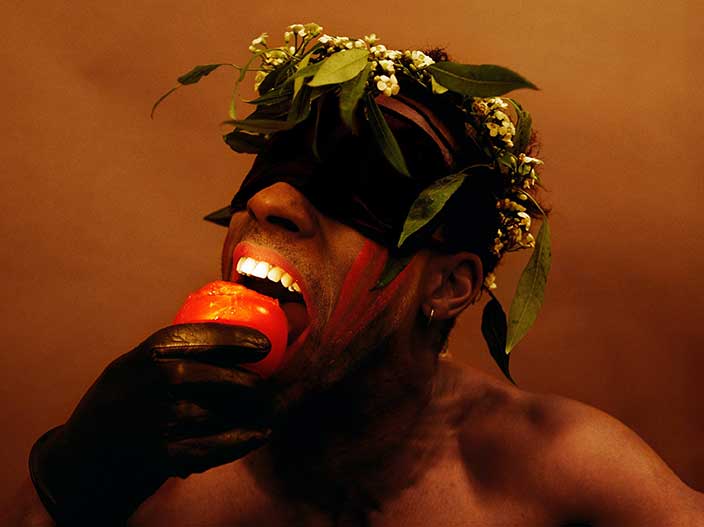 A photograph features a man with dark skin wearing a black blindfold and flower crown holding a red fruit in a hand with a black glove. The light is focused on the man’s mouth moving to bite the apple. 