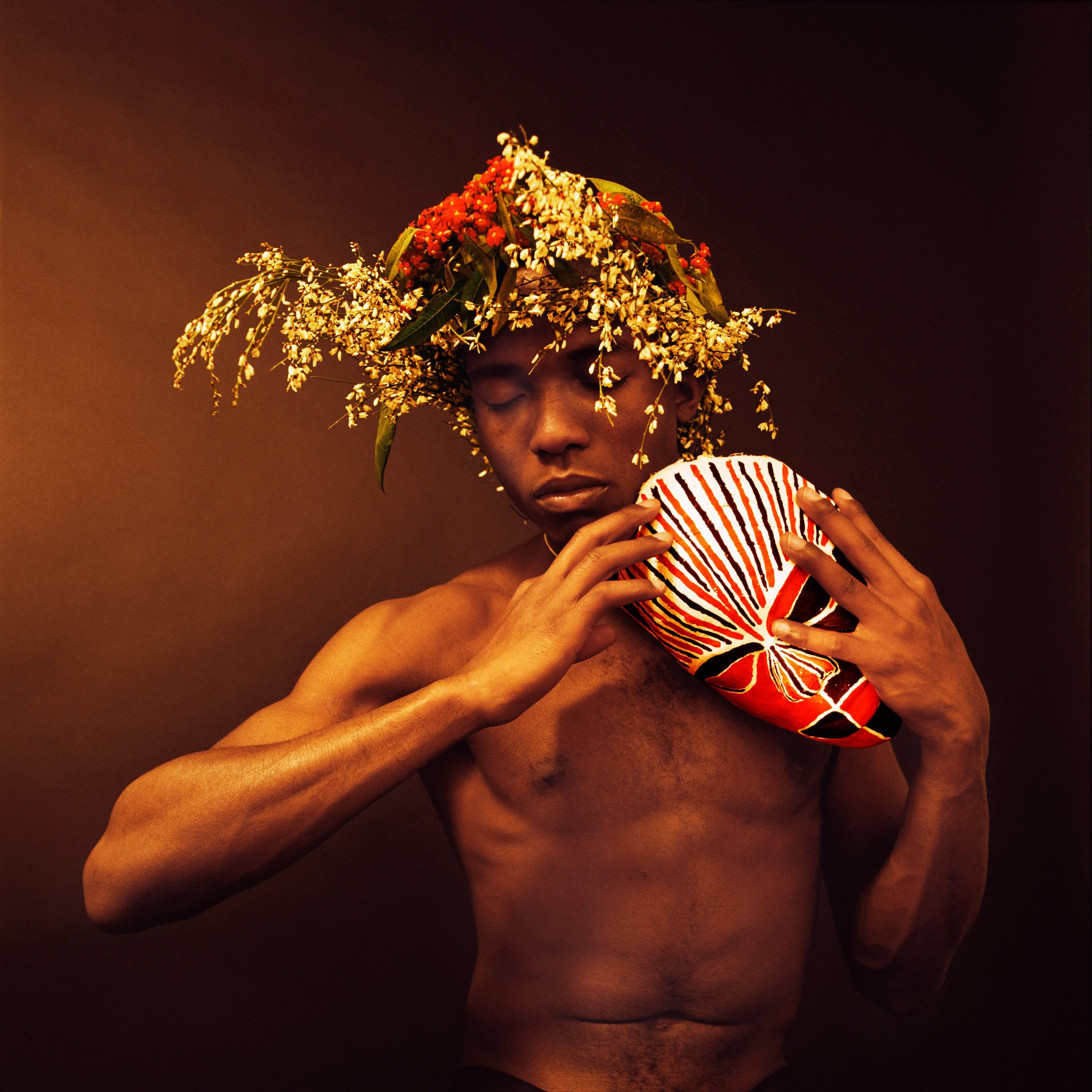 A photograph depicts a young man with dark skin wearing an elaborate floral head piece. He is shirtless and holding a red and black mask to his shoulder, his eyes closed.
