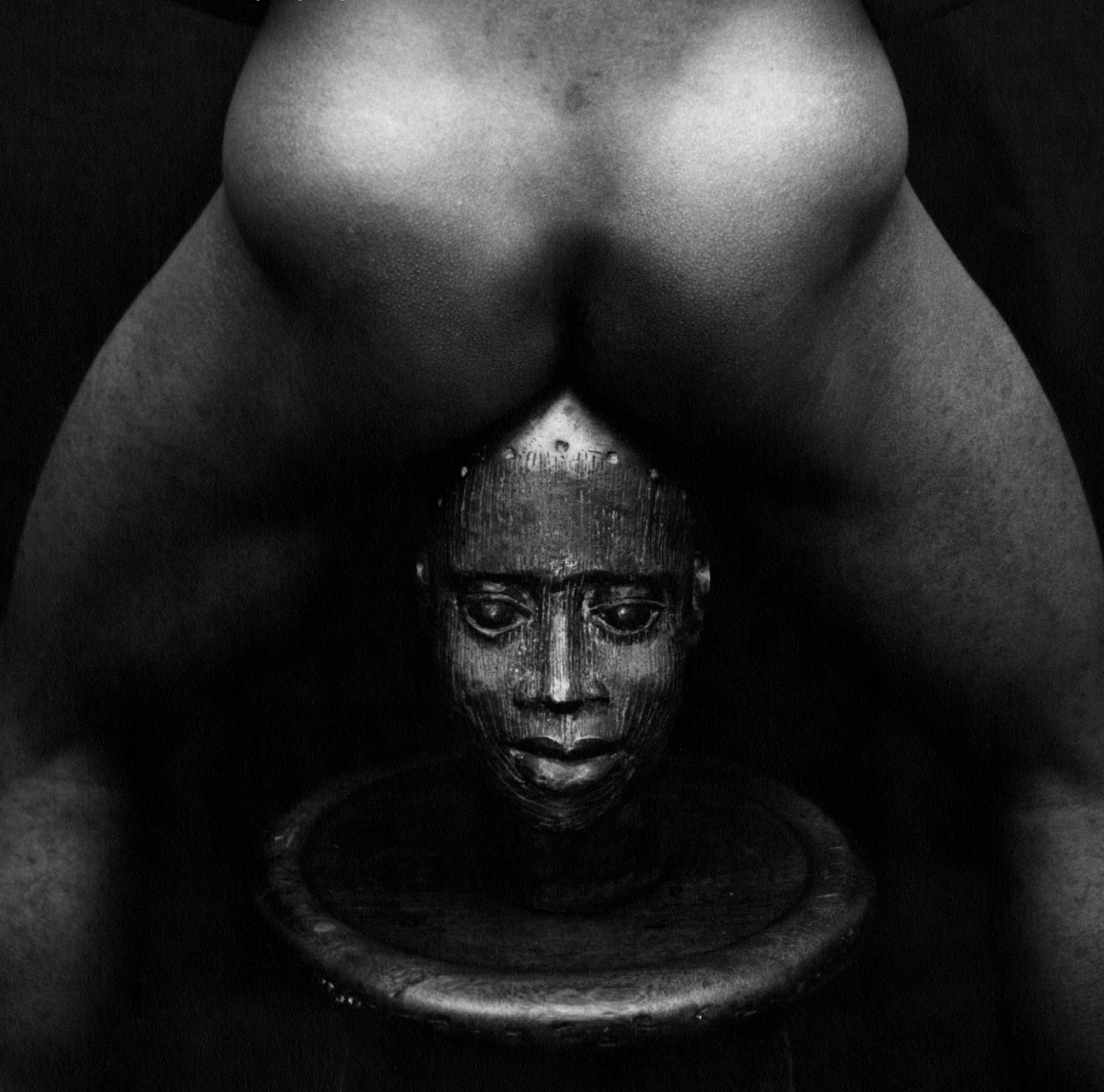 A black and white photograph depicts a man’s backside straddling a Yoruba mask.