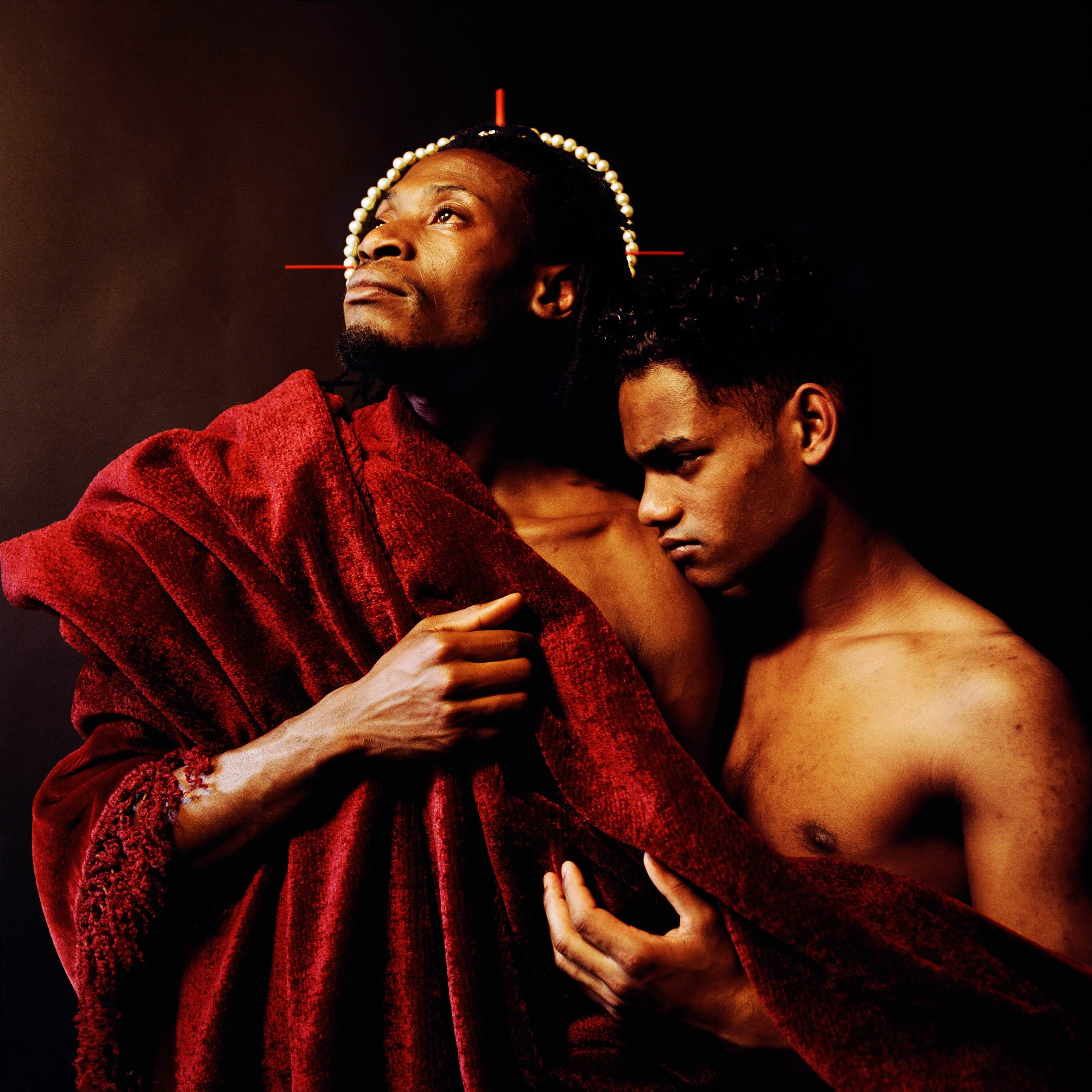 A photograph features two men posing together. The man on the left wears a red robe with a head piece evocative of religious imagery; the man on the right is shirtless and leaning on his shoulder.