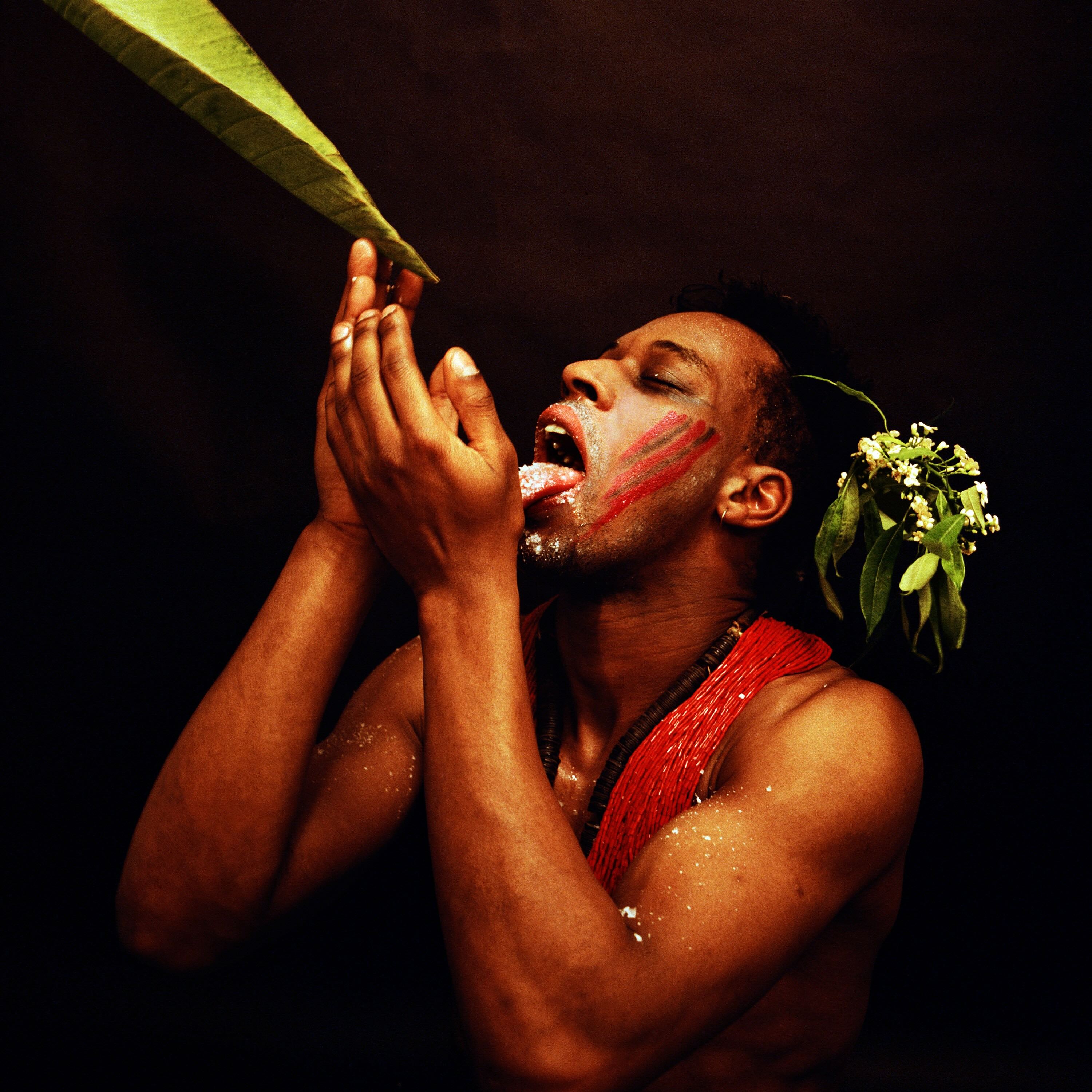 A photograph features a man with dark skin against a black background. He is reaching for a leaf and motioning as if to drink from it. He is wearing a blossoming leafy branch in his hair.