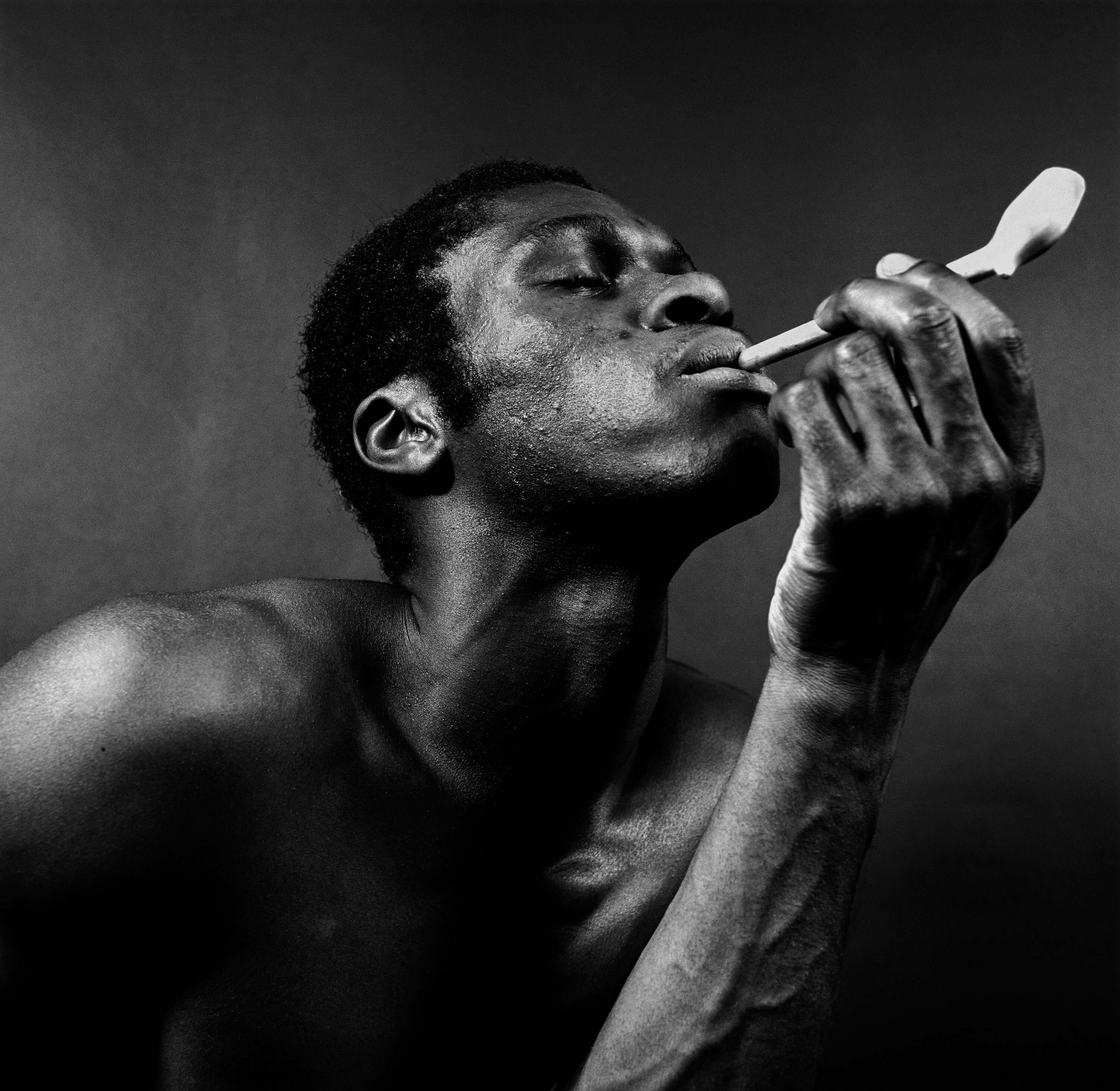 A black and white photograph features a man with dark skin holding a white pipe to his lips.