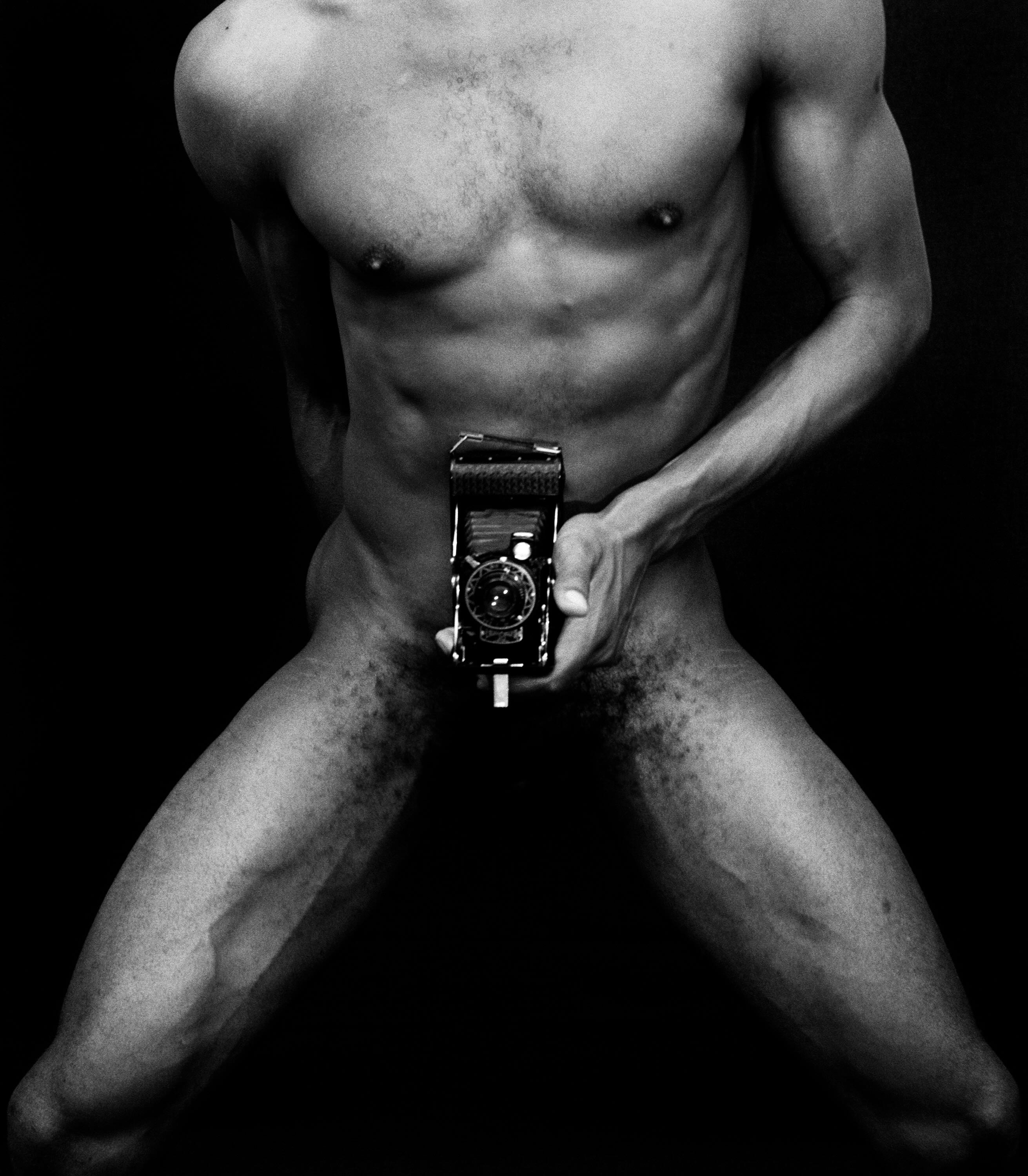 A black and white photograph features the torso of a man holding a camera near his groin.
